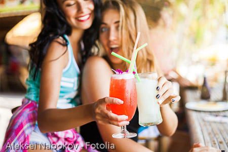 Two trendy cool hipster girls, friends, the blonde and the brunette drink cocktails at the cafe on the beach. Dressed in colorful shirts and shorts.